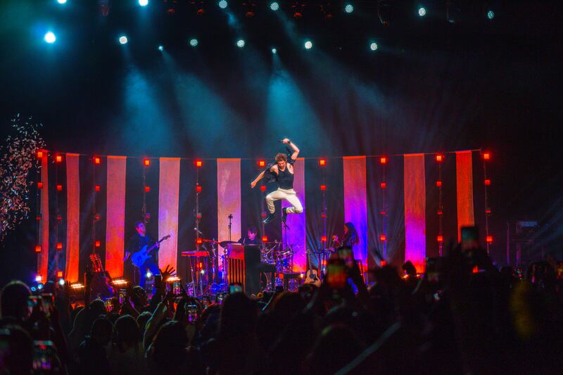 A wide shot photo of Bassett jumping up on stage. He is high up in the air and there are red lights behind him.