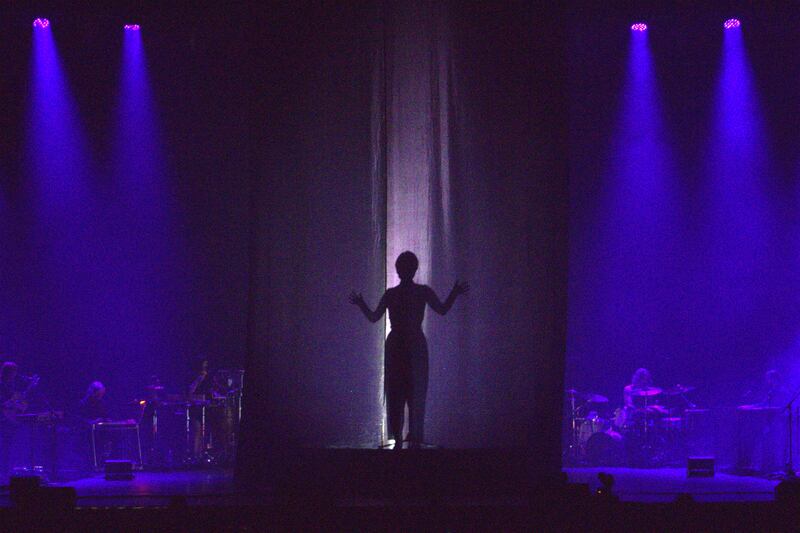 Photo of a woman's silhouette behind a curtain on a purple stage with a band flanking the curtain