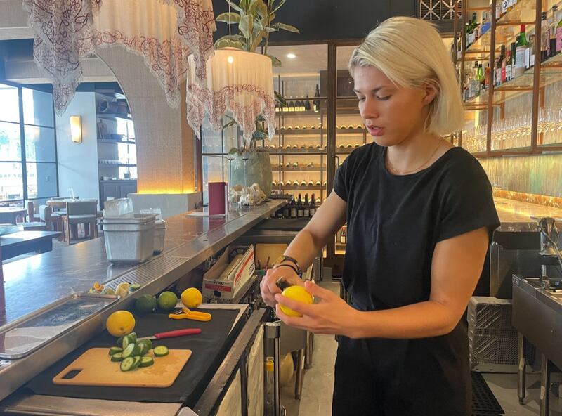A woman in a black shirt with icey blonde hair peels the rind of a lemon while behind the bar in a chic resturant