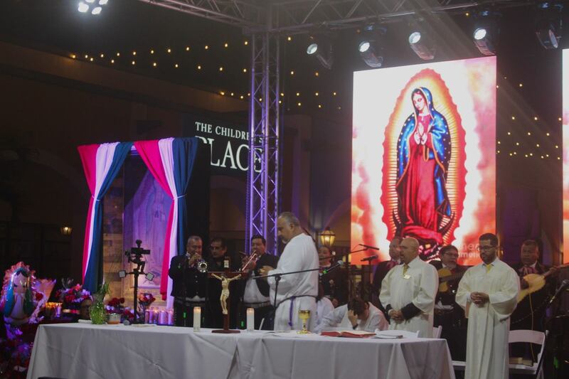 Photo of priests and musicians standing in front of an image of the Virgen de Guadalupe.