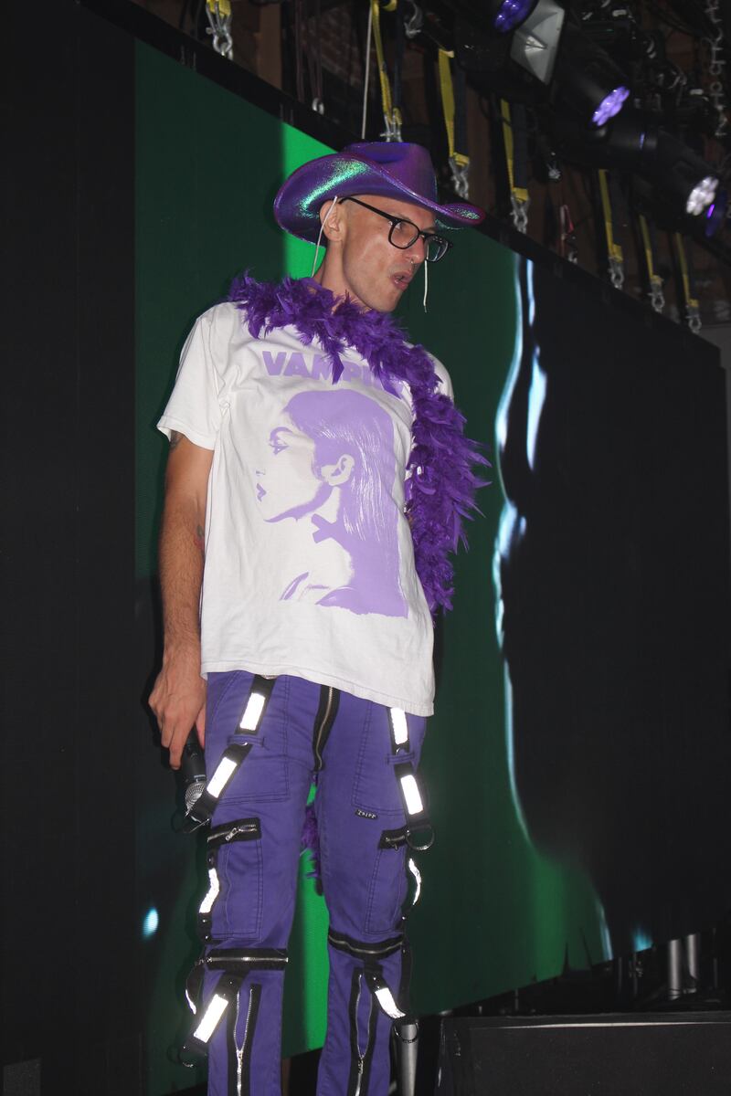 The DJ stands on stages in purple pants, a purple and white "Vampire" shirt, a purple boa and a purple cowboy hat.