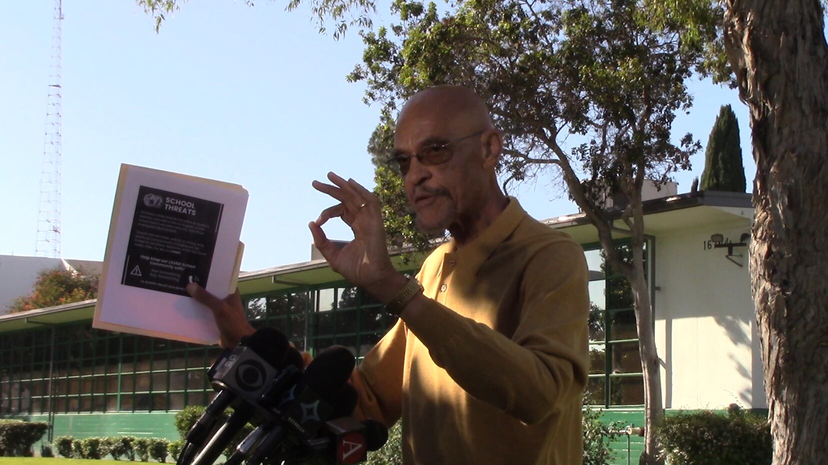 Person in a yellow shirt speaks at a podium with several microphones in front of a school, trees, and a blue sky.