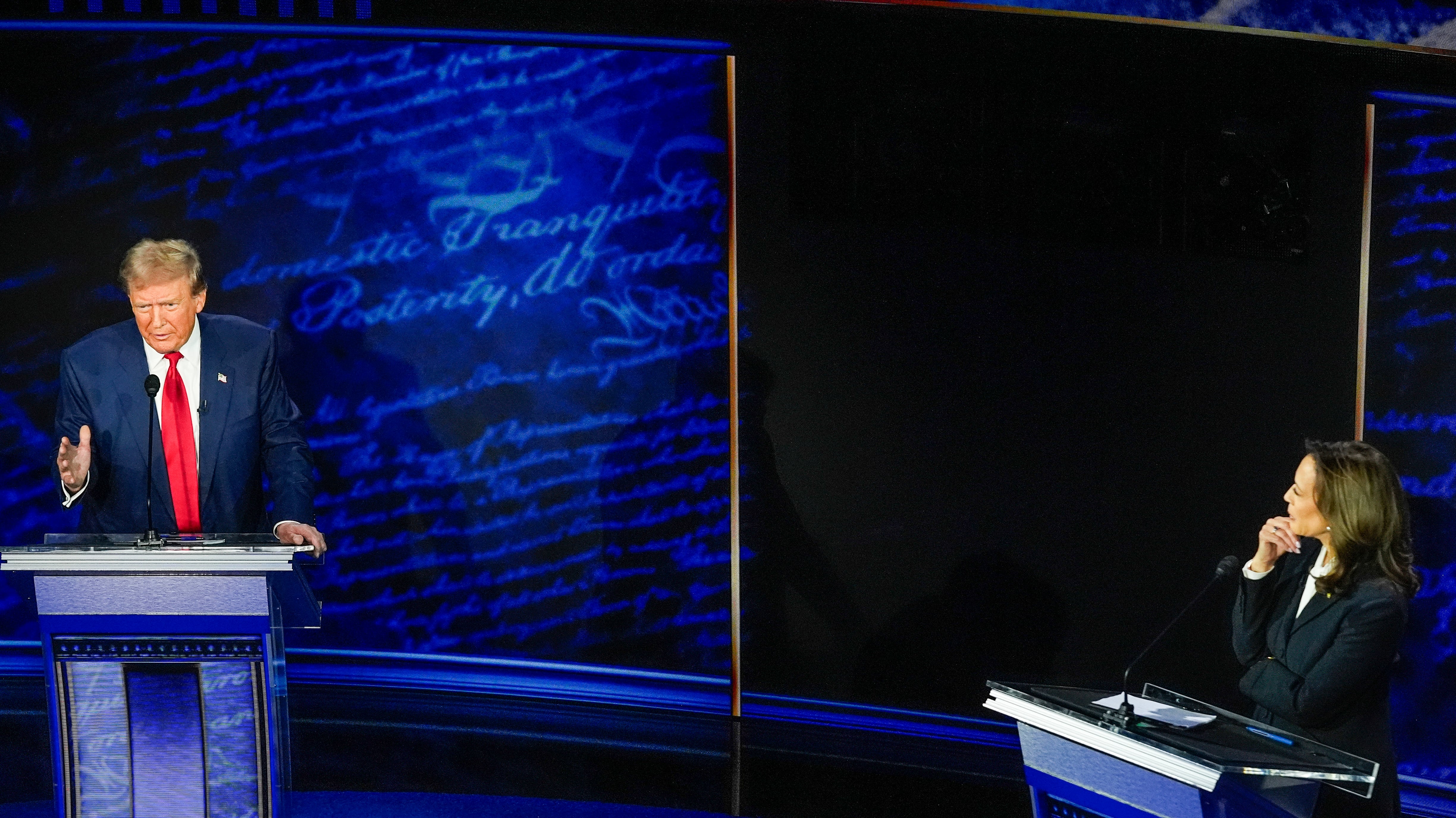 Man talking and women starting at him while standing on debate stage podium.