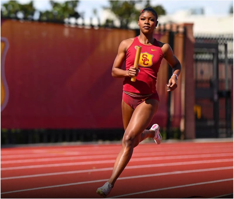 Photo of a Jassani Carter, golden baton in hand, running a relay race in her red USC track uniform
