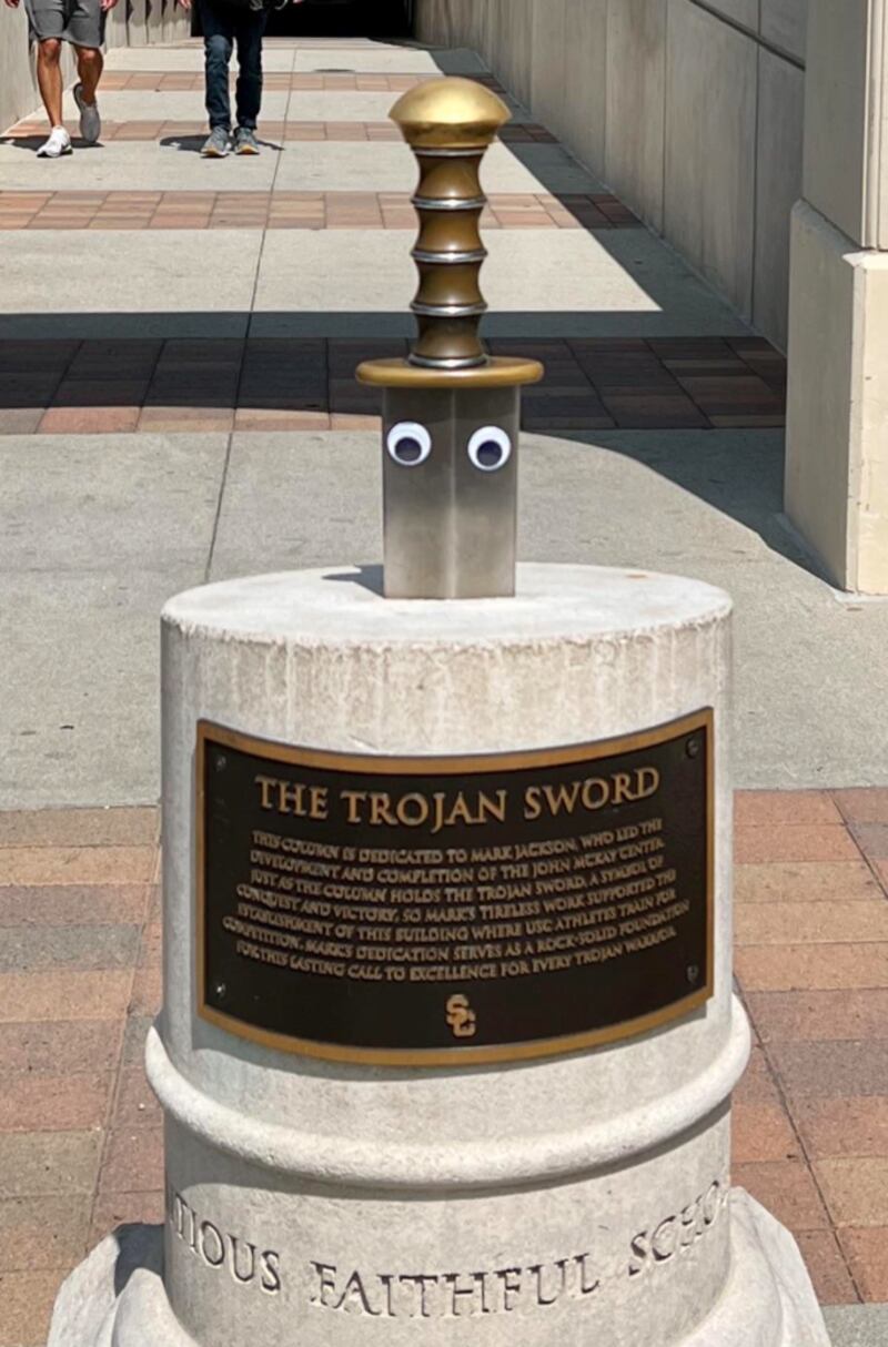 A stone podium with a metal plaque holds the sword, half of which protrudes. Google eyes sit on the blade of the sword. It is a sunny day and the path can be seen in the background.