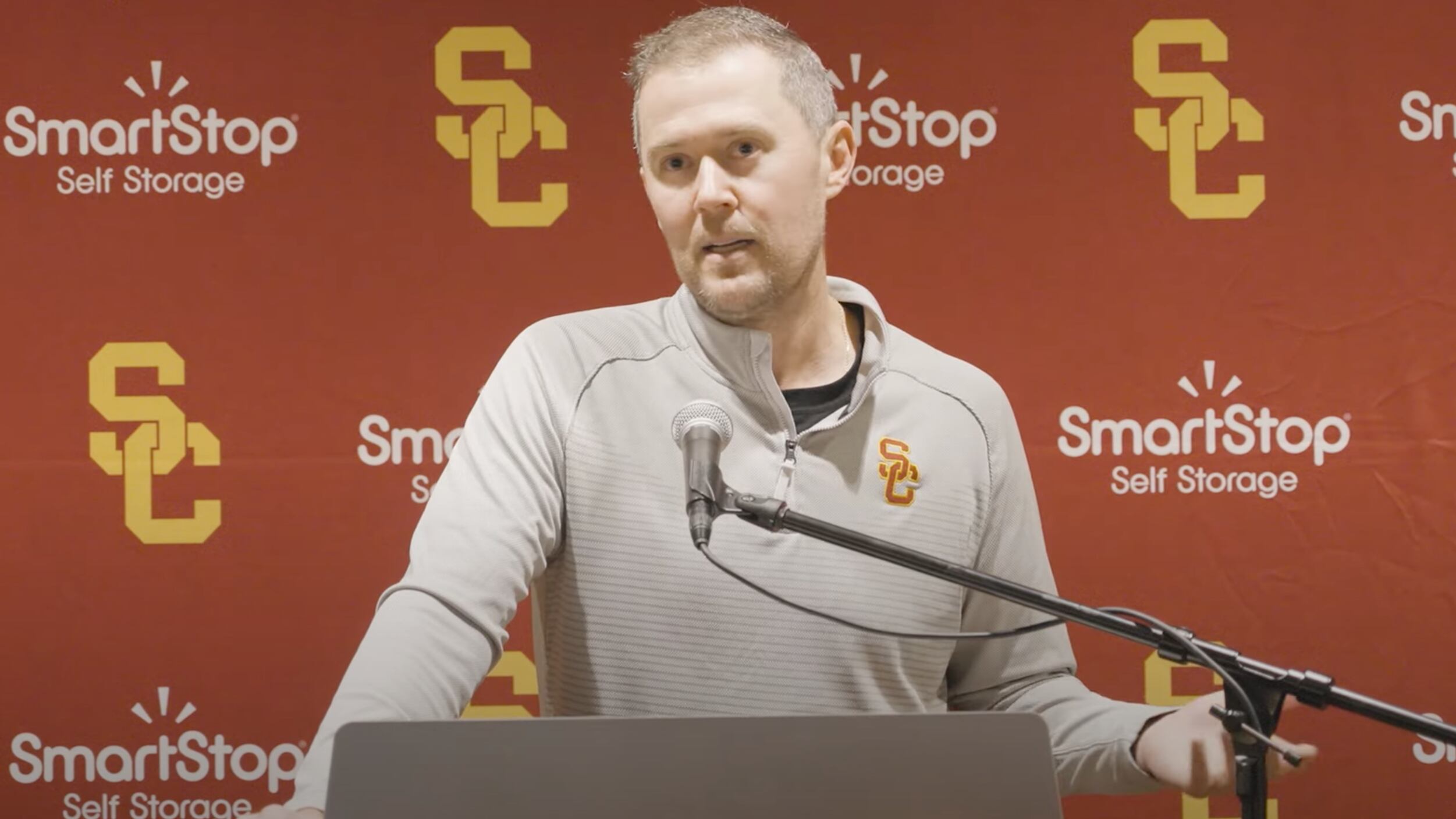 Lincoln Riley speaks at a pedestal in front of a step and repeat.