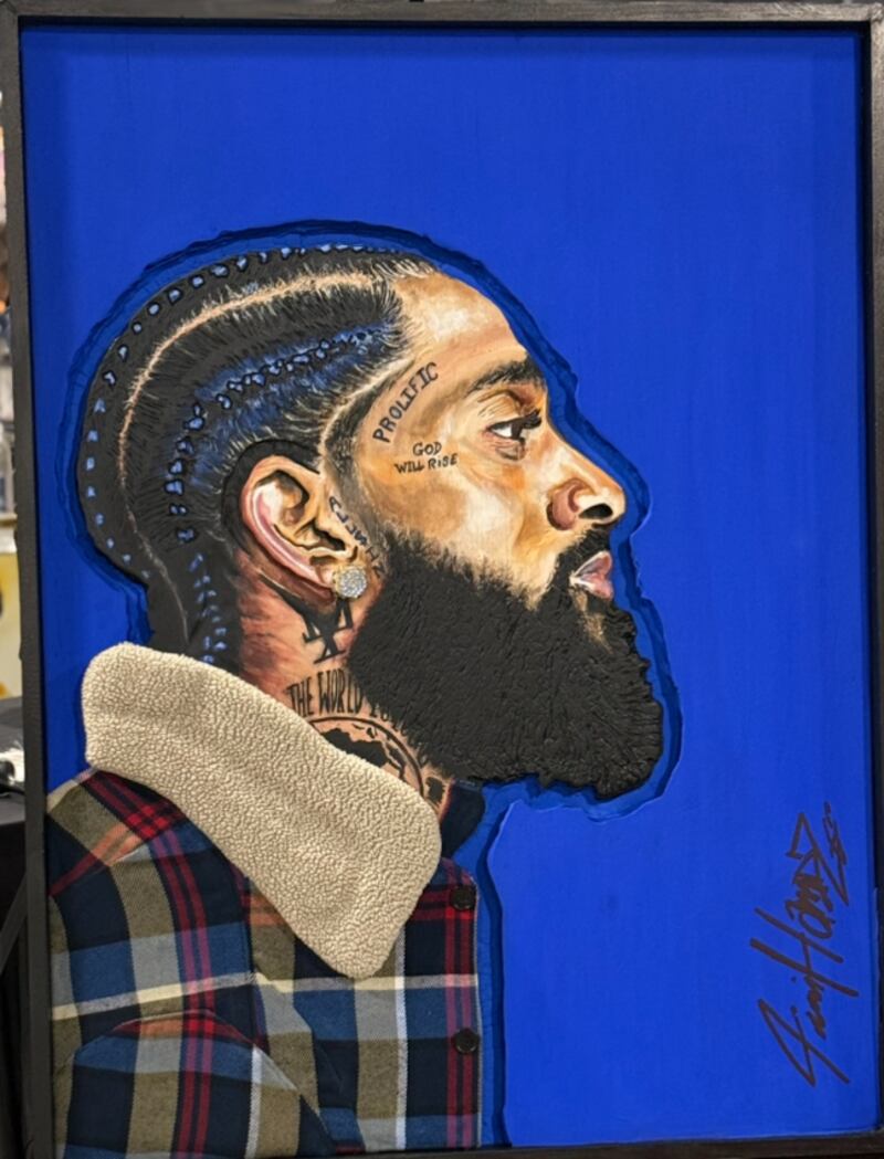 A framed portrait painting shows Nipsey Hussle's side profile with braided hair and a full beard, set against a solid blue background. He wears a plaid jacket with a shearling collar, and detailed tattoos are visible on his face and neck.