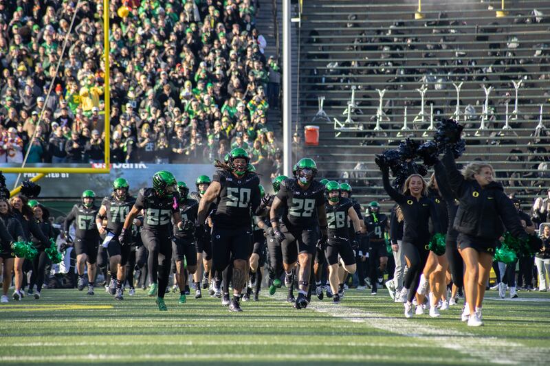 Oregon football players take the field at Autzen Stadium wearing black uniforms and green helmets.