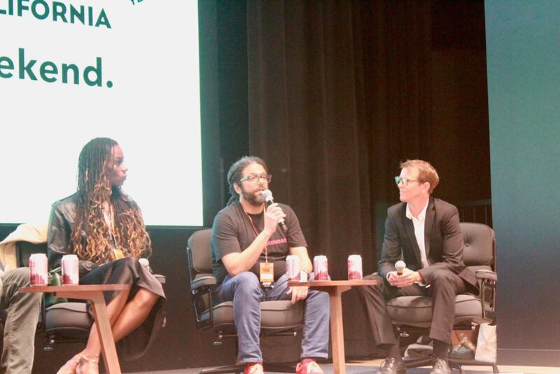 Frank is holding the microphone and speaking. He is wearing jeans and a black shirt. Both the panelist on his left and right and listening intently.