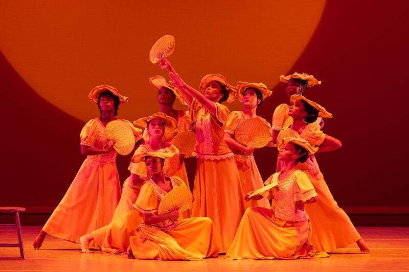 Dancers in flowing yellow dresses hold fans and pose together on a stage bathed in warm orange light, creating a unified and celebratory scene.