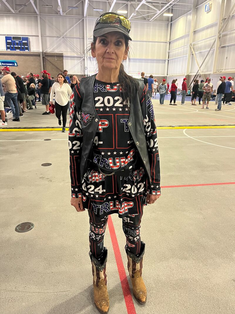 A woman wearing a baseball cap, sunglasses, a leather vest, cowboy boots, and a head-to-toe black "Trump 2024" outfit.