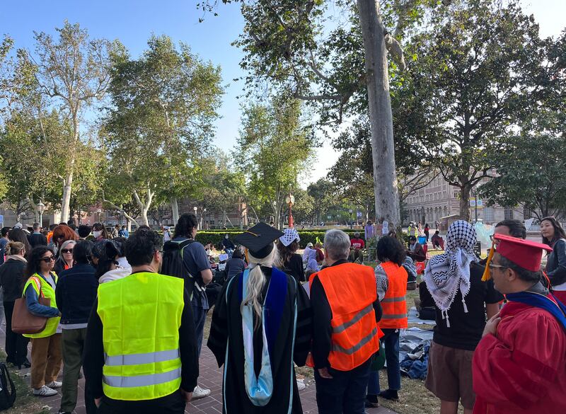 People in bright yellow and orange safety vests and academic regalia stand gathered together outside.