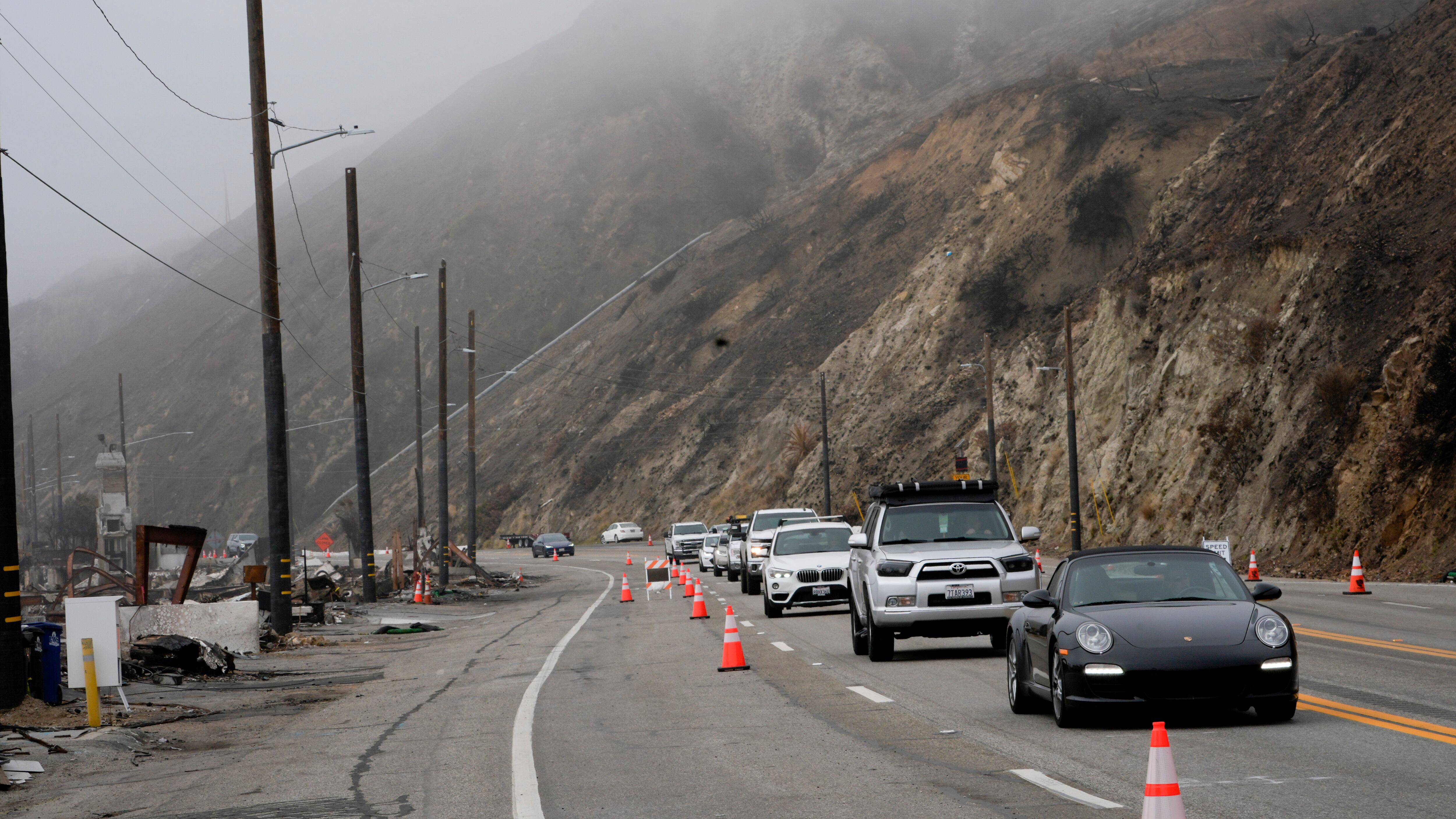 Cars drive on one side of the road, with the cliffs covered in fog and fire debris