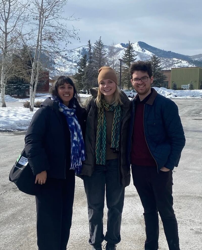 Three people in winter gear stand in a parking lot, with a snowy mountain behind them.
