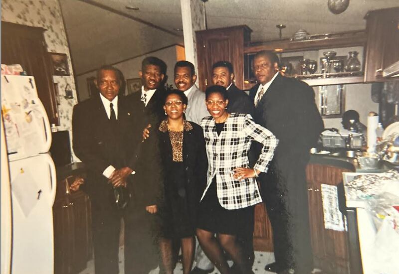 A group family photo taken indoors, showing several adults dressed formally and standing together in a kitchen. Brenda Smith stands in the front row on the right, smiling, with other family members gathered around her.