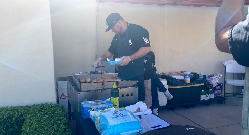 Joh is wearing a black hat and shirt with a chefs apron on. He is cooking burger buns on a portable grill outside of the building.