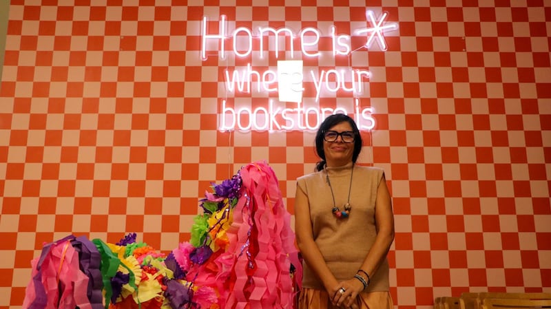 Photo of a woman standing in front of a pink and red checkered background and a neon light that reads 'Home is where your bookstore is.'