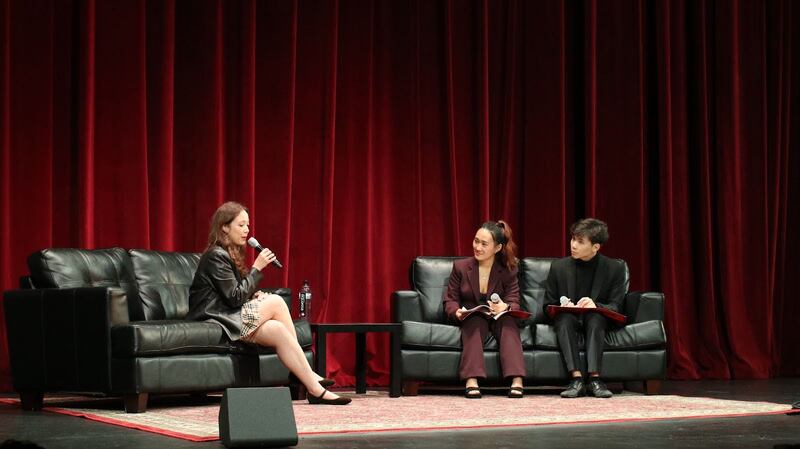 Laufey sits on a leather couch in a skirt and leather jacket speaking into a microphone. Two moderators (one in a maroon suit and one in black suit) sit across from her on a leather couch.