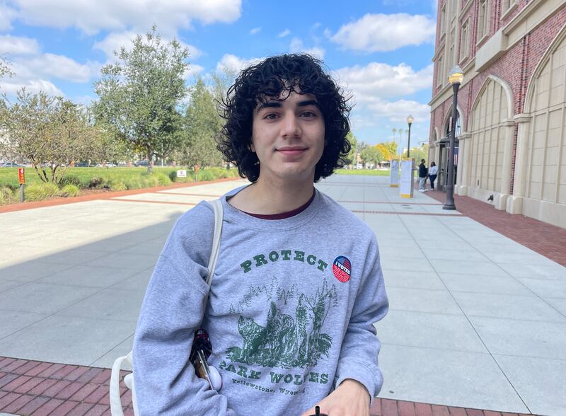 Serrano stands between a lawn and a brick building, wearing an "I Voted" sticker.