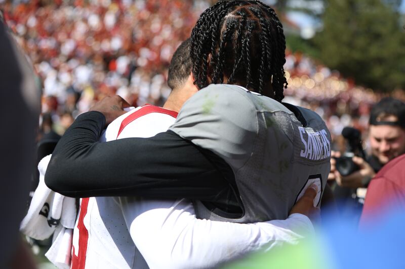 Caleb Williams and Shedeur Sanders hug after a USC/Colorado football game. Williams wears w hite football jersey with cardinal and gold accents, while Sanders wears a gray jersey with black and white accents.