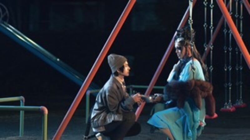 Film still of a person kneeling in front of another in a costume sitting on a swing