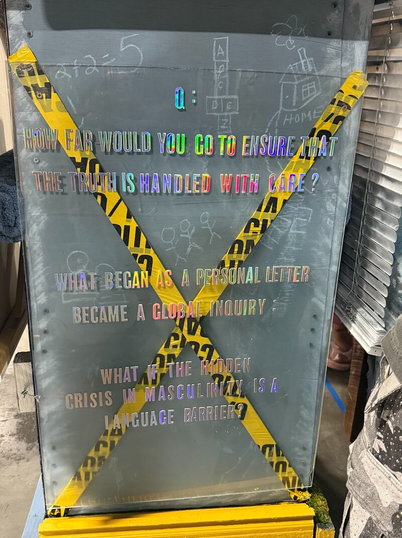 A mixed media text artwork features reflective lettering over a gray surface marked with chalk drawings and yellow caution tape arranged in an X shape. The text asks questions about truth, language, and masculinity, displayed inside an indoor exhibition space.