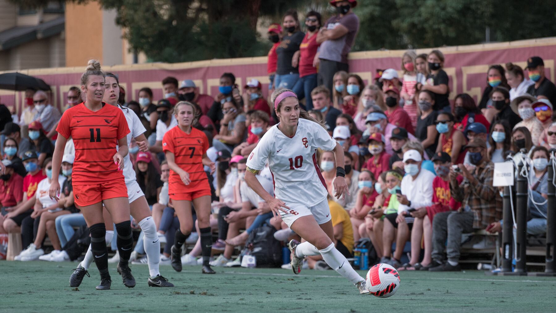 A photo of Savannah DeMelo in a white uniform dribbling a white and orange soccer ball. Oregon State players in orange uniforms are in the background, as well as a crowd of USC fans.