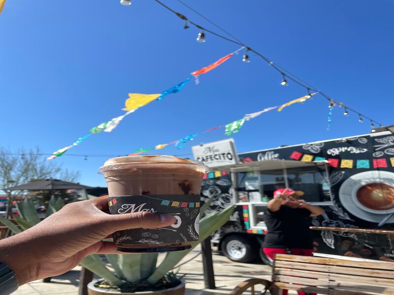 Photo of a hand holding a coffee with a person and trailer in the background.