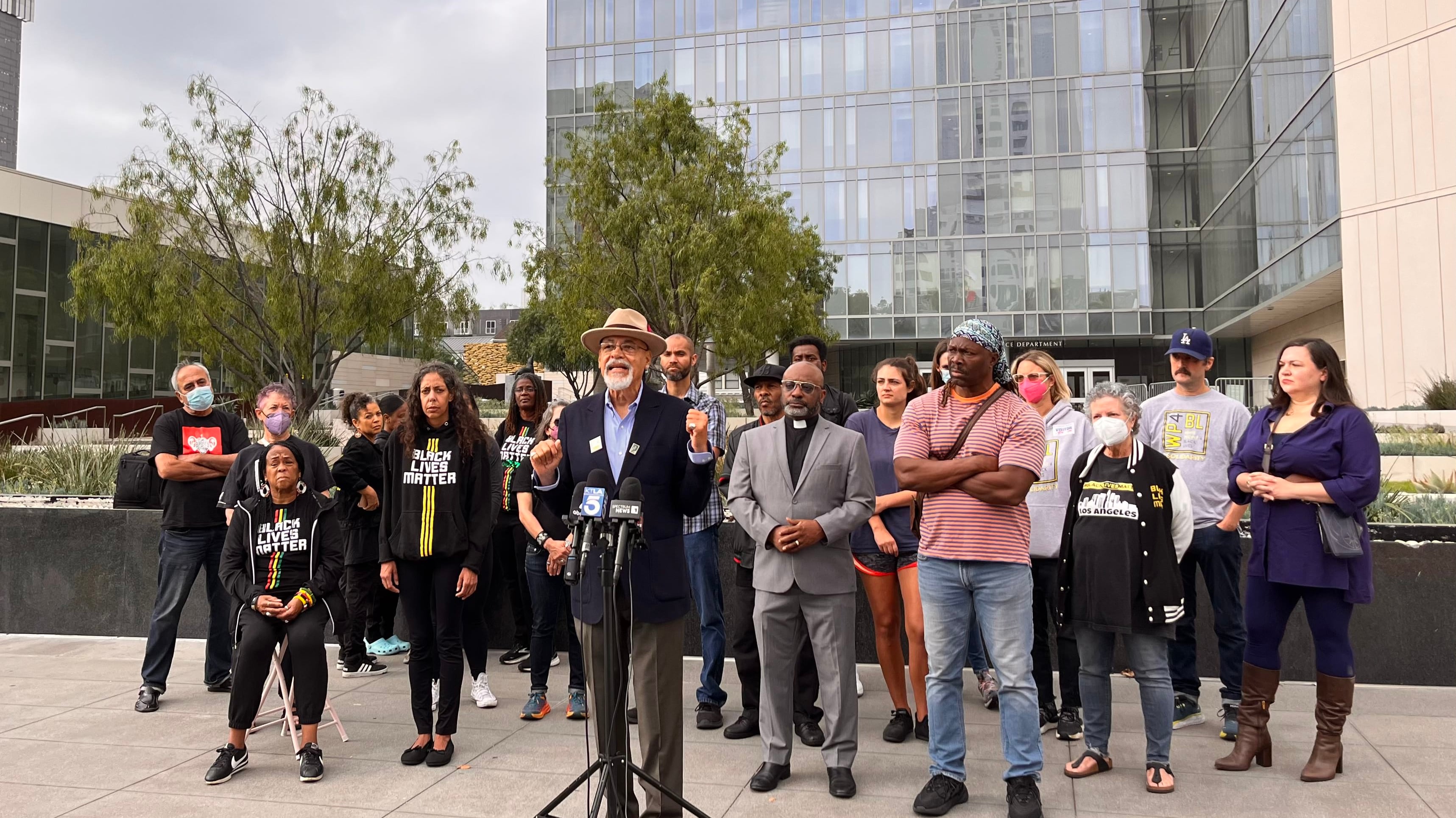 Greg Akili stands in front of a podium, surrounded by nearly 20 other activists, outside of the Los Angeles Police Department.