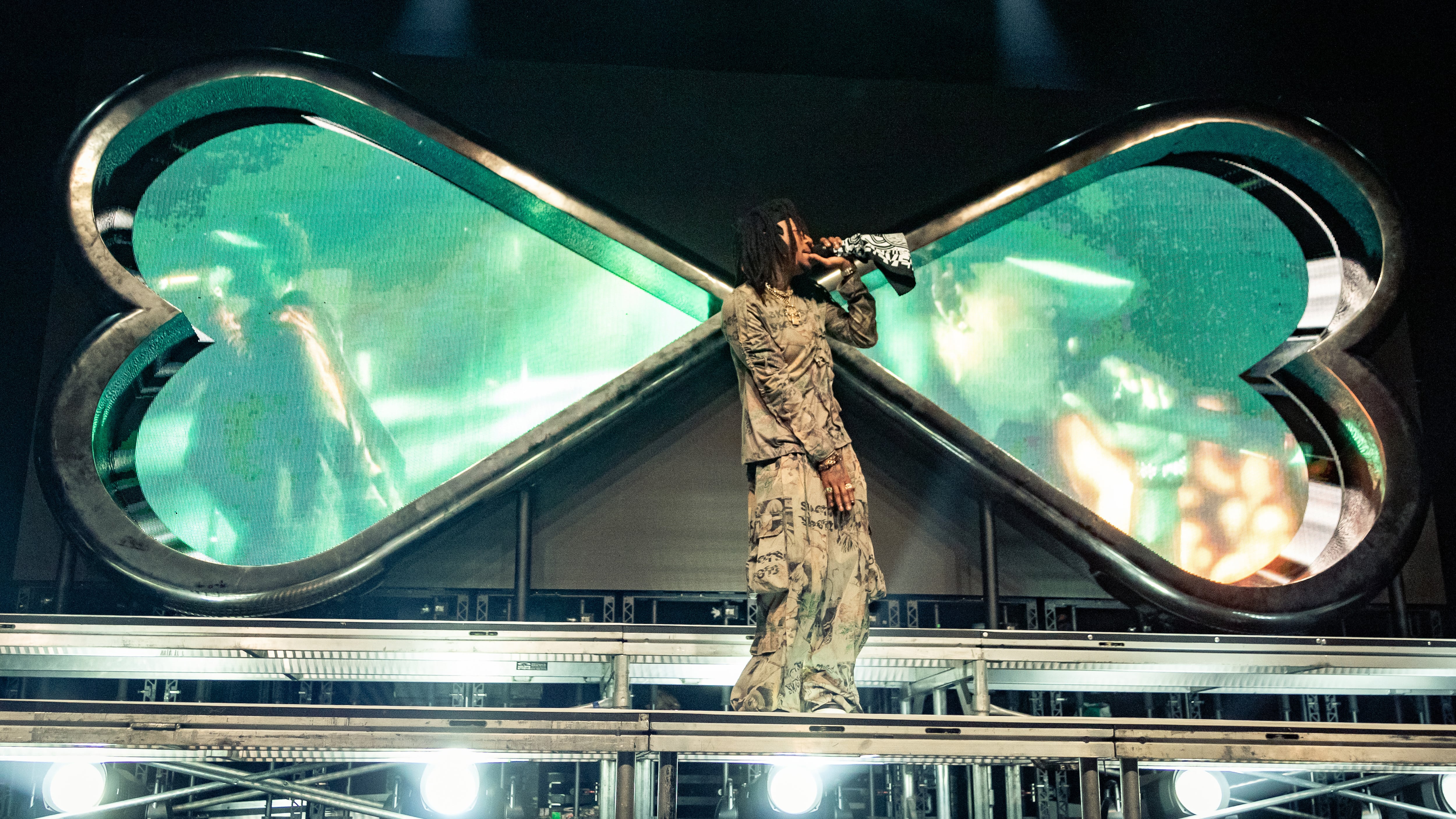 A photo of J.I.D. On the stage with big blue/green sideways hearts behind him in the set design.