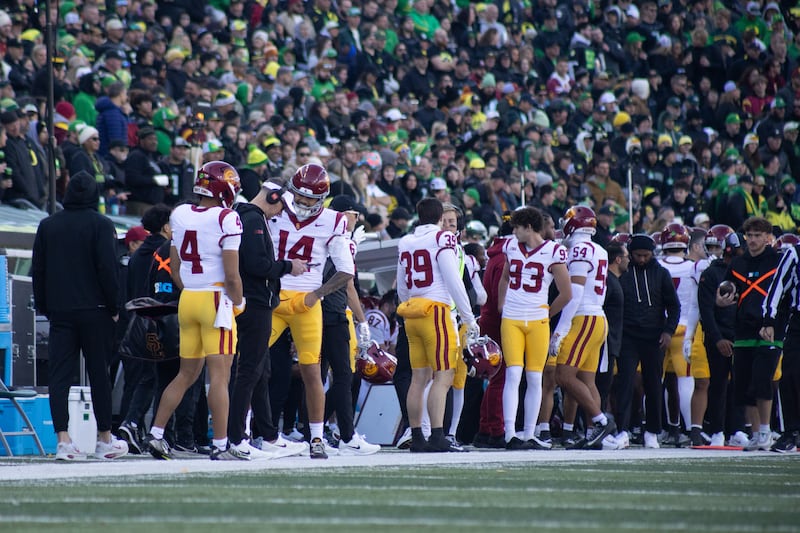 Jayden Maiava (14) and other football players on USC sideline. Maiava is in conversation with head coach Lincoln Riley, who wears a headset and looks at a tablet in his hands.