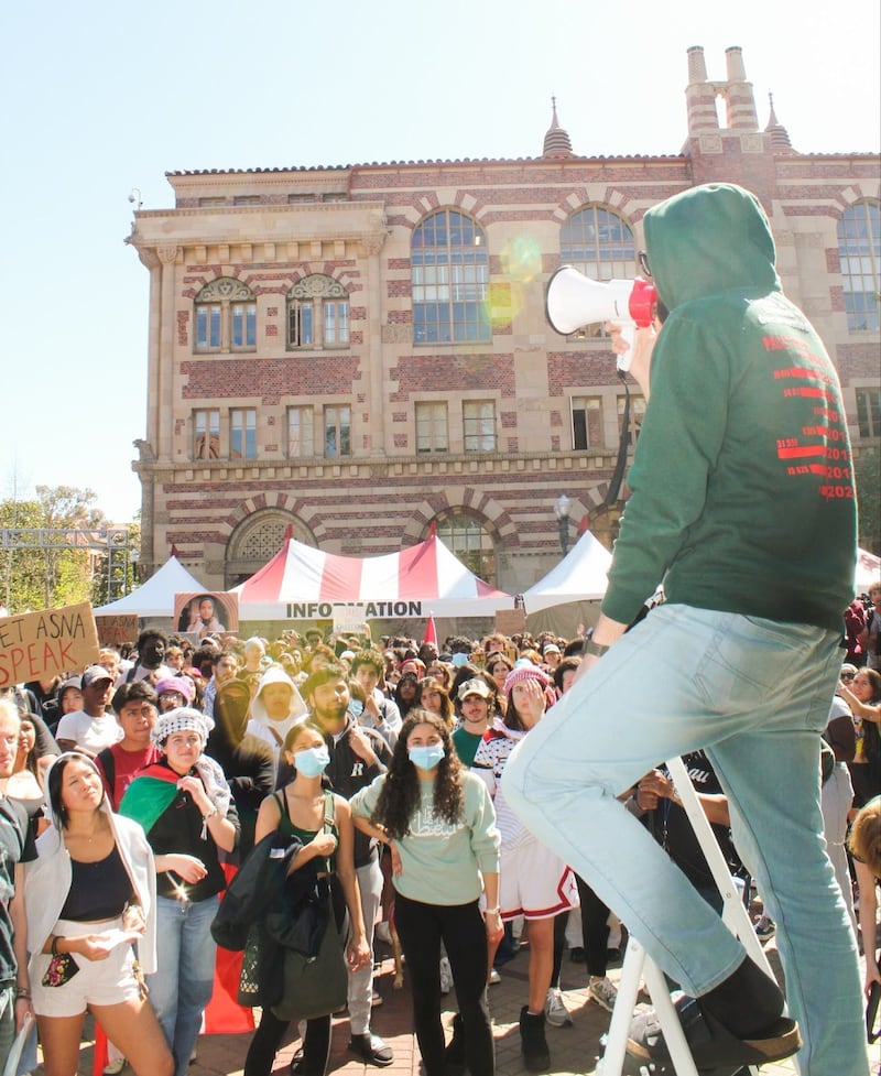 A man in a green hoodie stands at the top of a white step ladder with a megaphone in his hand. In front of him are hundreds of protestors, many masked, listening to his speech outside of a large brick and sandstone-colored building.