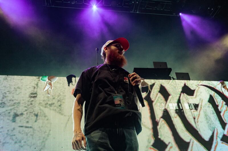 A photo of Freddie Dredd performing on stage and singing into a microphone. He's wearing a baseball cap, T-shirt, sunglasses and jeans/
