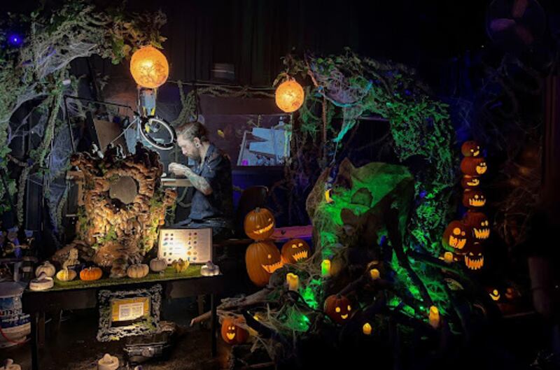 A jewelry-maker stands in a corner under a ring light, surrounded by Halloween decor such as a stack of plastic jack-o-lanterns.