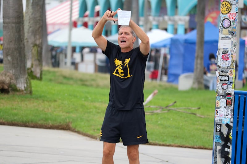 Eric Musselman stands with arms in the air, forming a diamond with his hands. He holds a pen and piece of paper and wears black USC basketball gear.