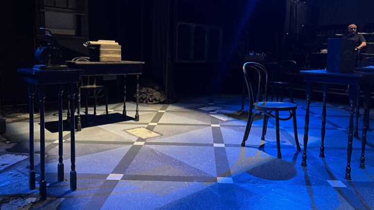 A small, dimly lit stage is adorned with basic chairs and desks. There is one larger desk in the center. The performance has not started.