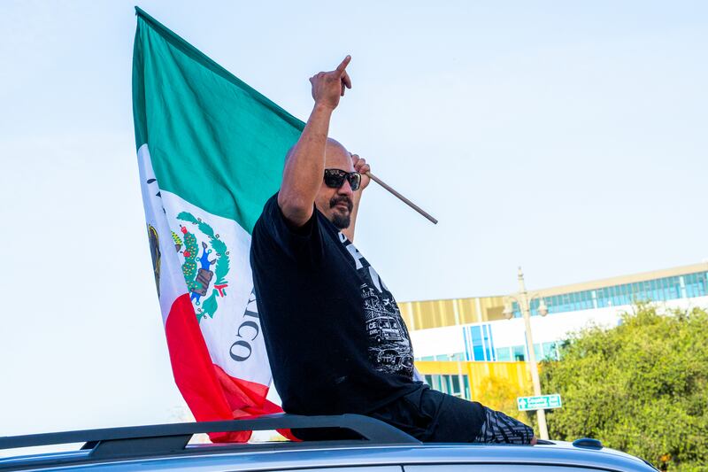 A man in sunglasses holds up his right hand, with his thumb and pinky extended from a fist, and sits atop the car, a Mexican flag in his left hand.