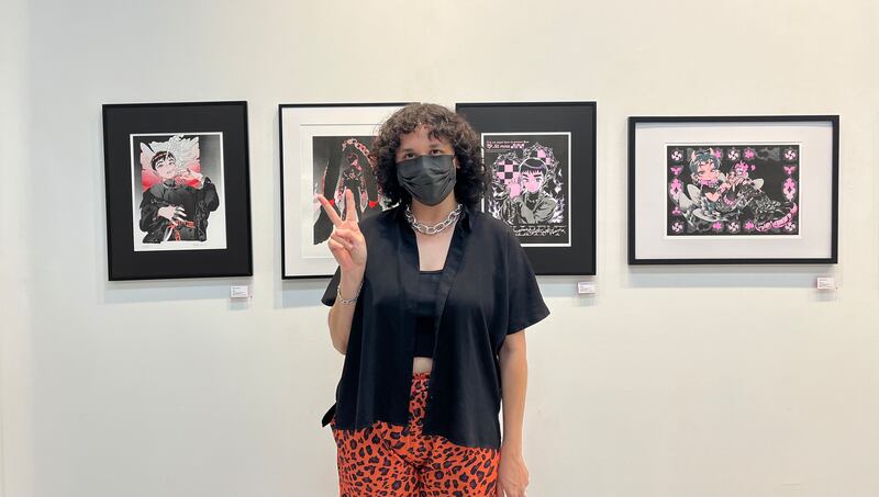 Photo of a person with a red shirt and leopard-print pants. They also wear a chain necklace and a black face mask. They stand in front of hung artwork