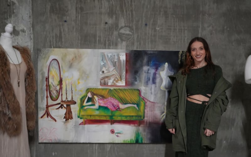 Berkowitz is wearing a black top and pants with a dark green coast around her arms. She is smiling at the camera and standing next to her painting. The painting has a woman in pink on a green couch.