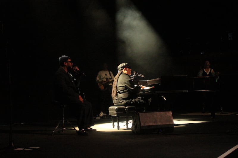 Stevie Wonder performs on the piano on a dimly lit stage, with the spotlight on him. PJ Morton sings next to him on a stool.