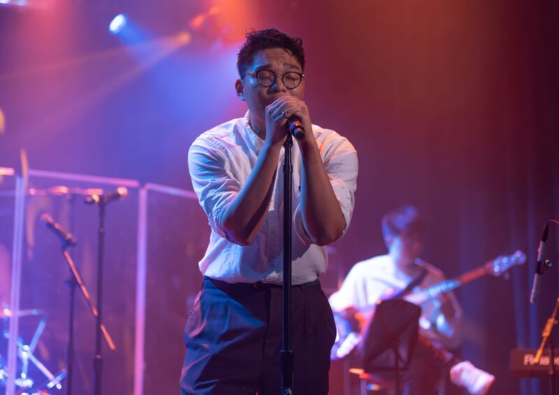 Photo of a man in glasses and a white shirt singing into a microphone on stage