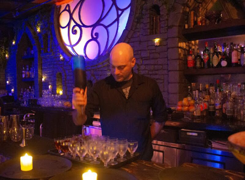 A bartender makes batch cocktails, holding a bottle upside down over the glasses.