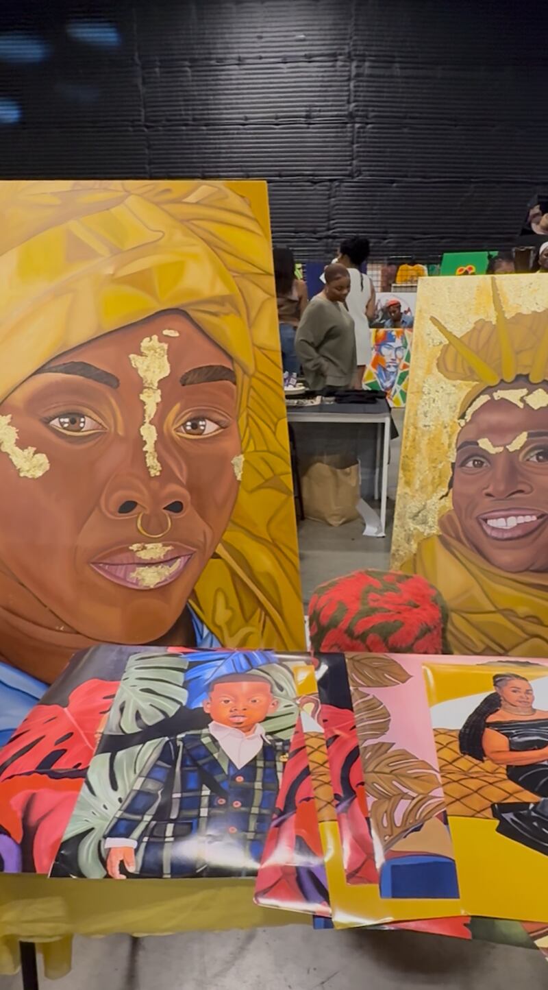 Two large portrait paintings of Black women wearing yellow headwraps stand behind a table covered with art prints. Smaller prints of children and women are arranged in front, while people move through the indoor event space in the background.