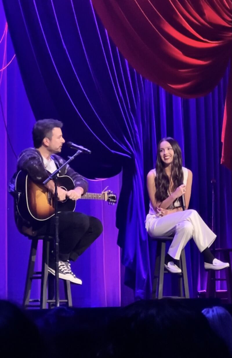 Olivia Rodrigo holds a microphone while looking at Dan Nigro speaking and holding his guitar.