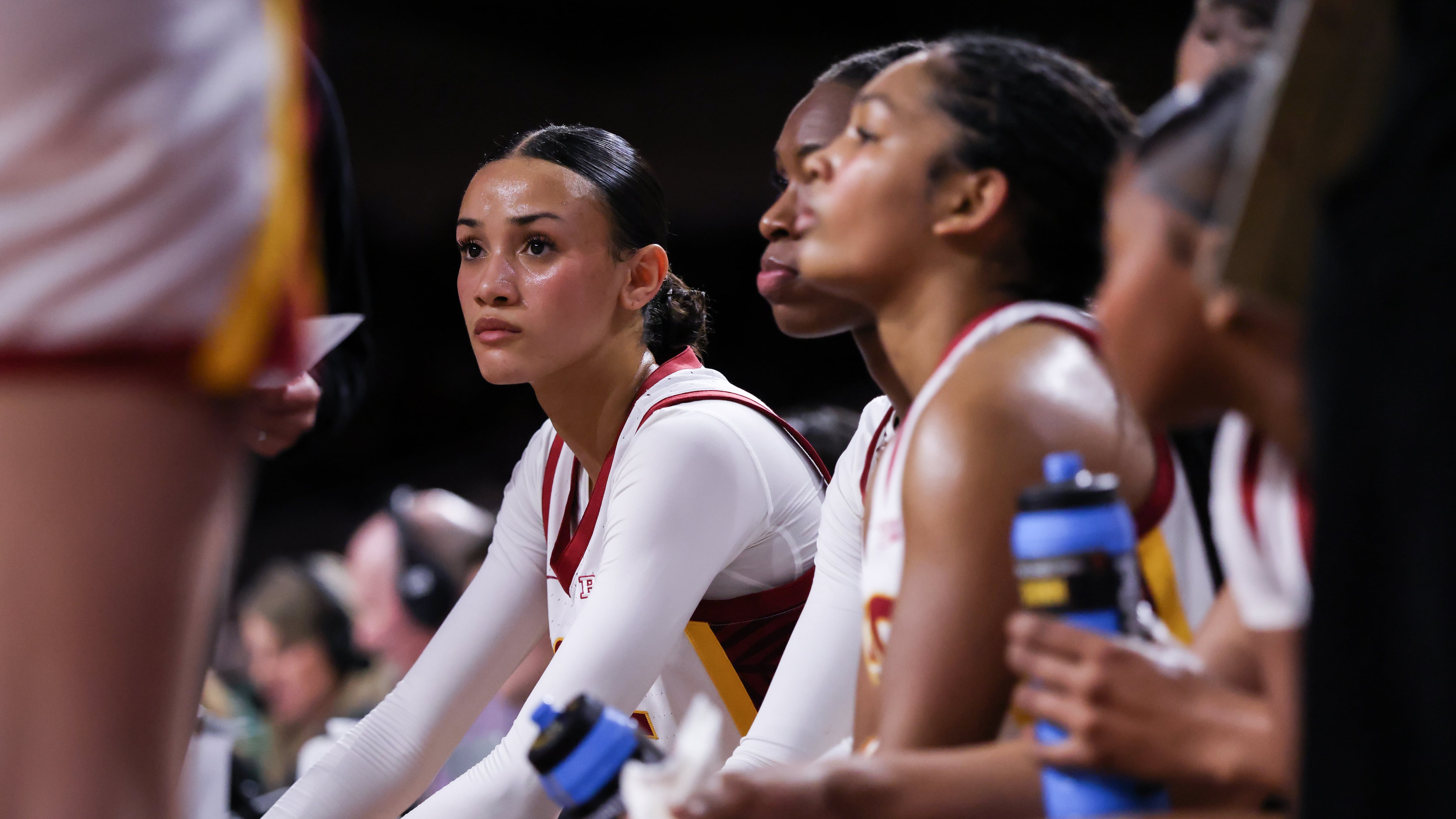 A female basketball player in a white uniform listens intently during a group huddle.