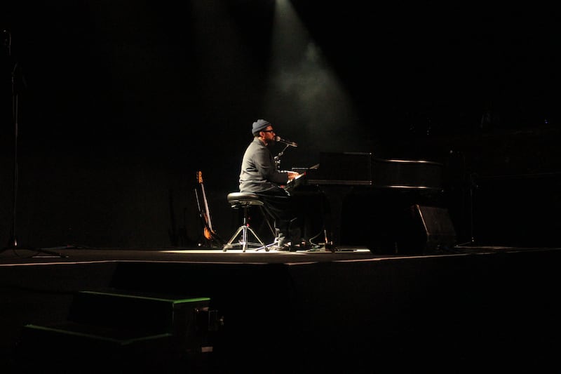 PJ Morton plays the piano alone on a dimly lit stage. The spotlight is on him and he is wearing all-black clothes.