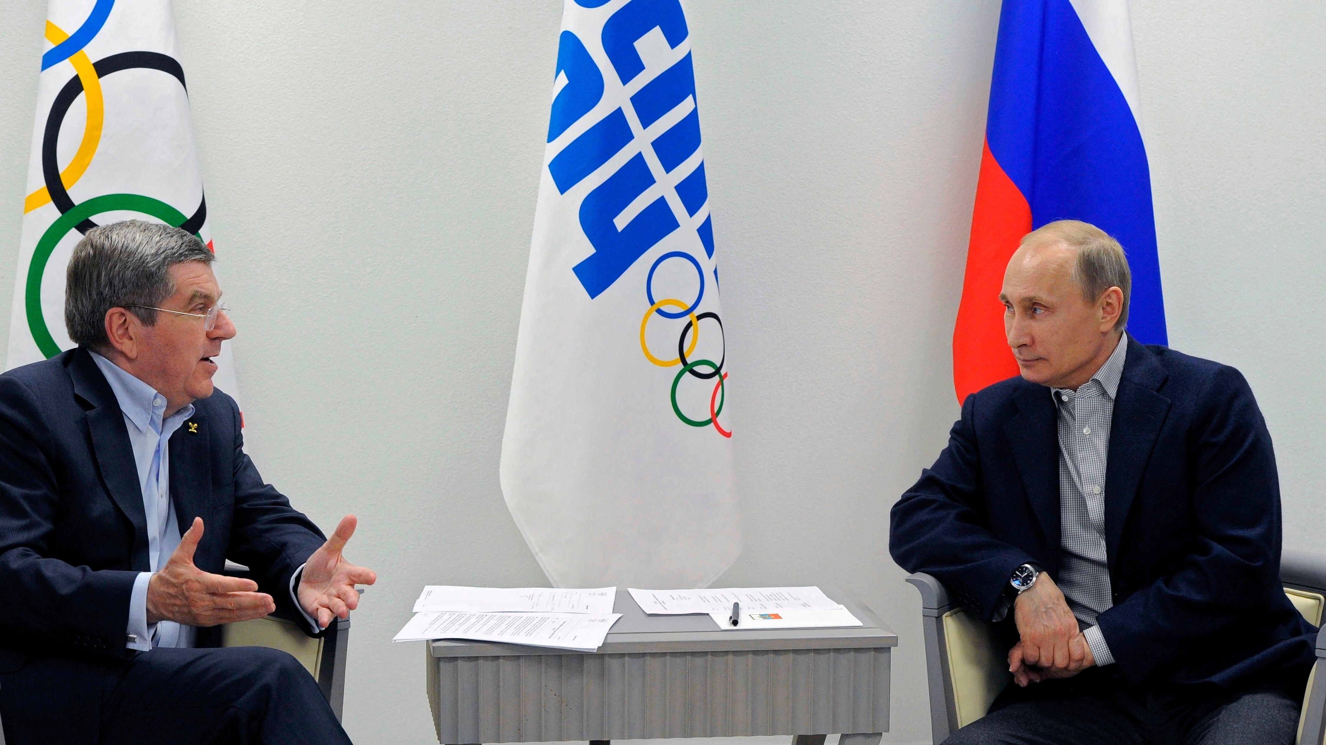 Putin is meeting with the IOC President Bach. They are sitting down and both in black suits with a Russian flag and Olympic flag behind them.