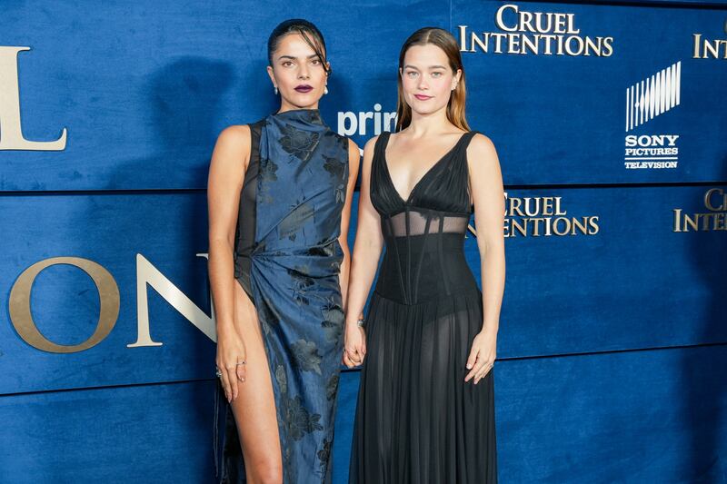 The two pose on the blue carpet. Silva is wearing a blue dress with black flowers and a slit and her hair is in a slicked-back poney tail. Hook is wearing a long black dress and her hair is down and tucked behind her ears.