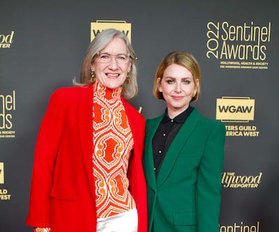 A photo of Henson (in an orange top and suit jacket) and Feldman (in a black blouse and green suit) smiling and posing on the red carpet.