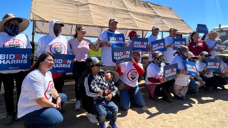 Over a dozen UFW members and their family members pose with Biden-Harris shirts and signs on a dusty patch of dirt between cultivated fields. (Photo by Jules Feeney)